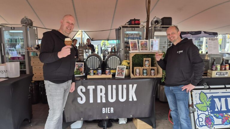 Twee mannen in Struuk Bier-hoodies proosten met een glas bier achter hun stand met tapvaten, blikjes en posters onder een tent