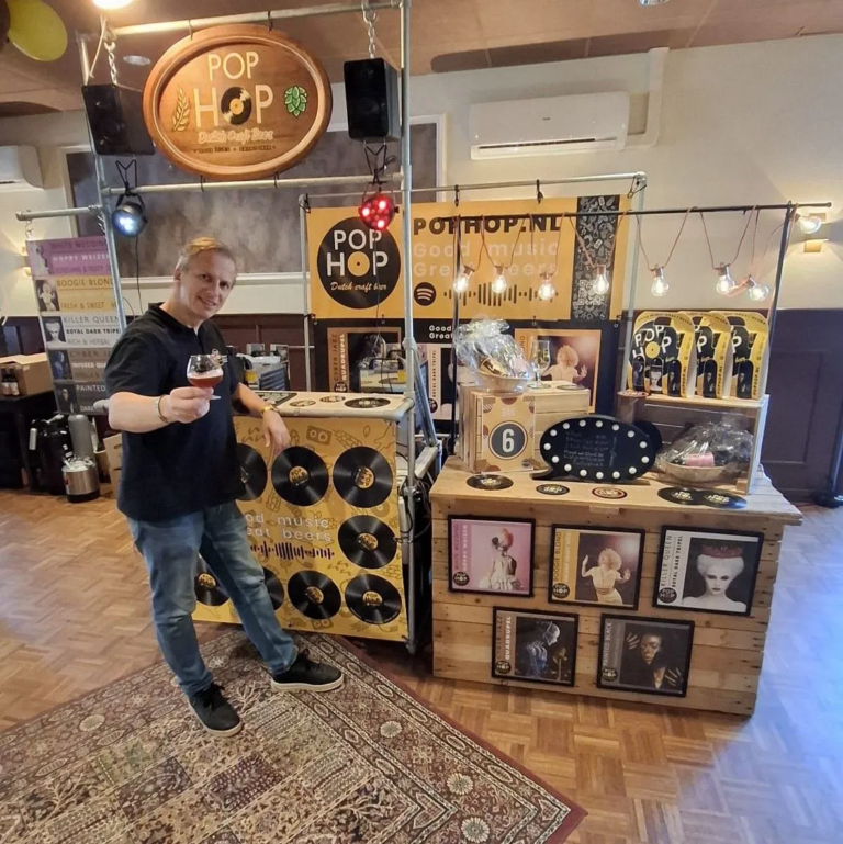 Man met een glas bier bij de PopHop-stand op een festival, met platenhoezen en vinylplaten als decoratie