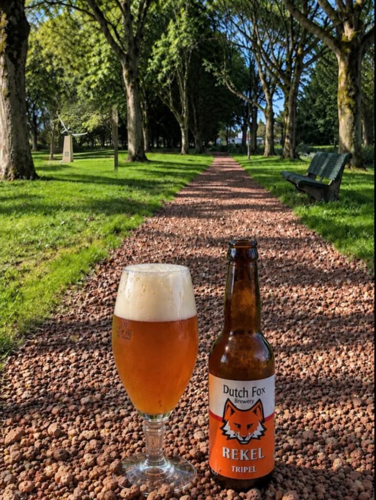 Rekel Tripel van Dutch Fox, glas en fles op een rood grindpad in een bomenlaan met een bankje verderop