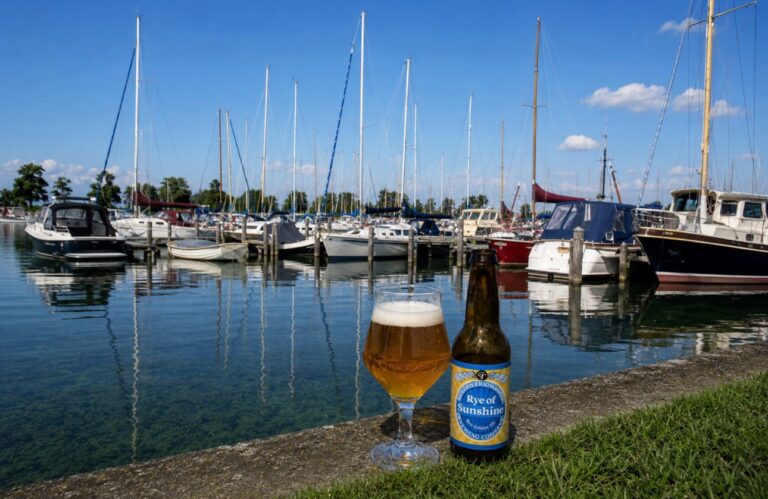 Rye of Sunshine van Gulden Triomfator, glas en fles op de kade van een jachthaven met zeilboten