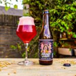 Sauwelaar Braam en Framboos van Het Zwijn, robijnrood glas en fles op een houten tafel met mout in een groene tuin