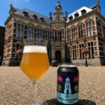 Hoppy Invaders NEIPA van UTCA, glas en blik op een plein voor een monumentaal bakstenen gebouw in de zon